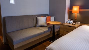 A hotel room with a grey sofa, a small round table, a bedside table with a lamp, and a bed.