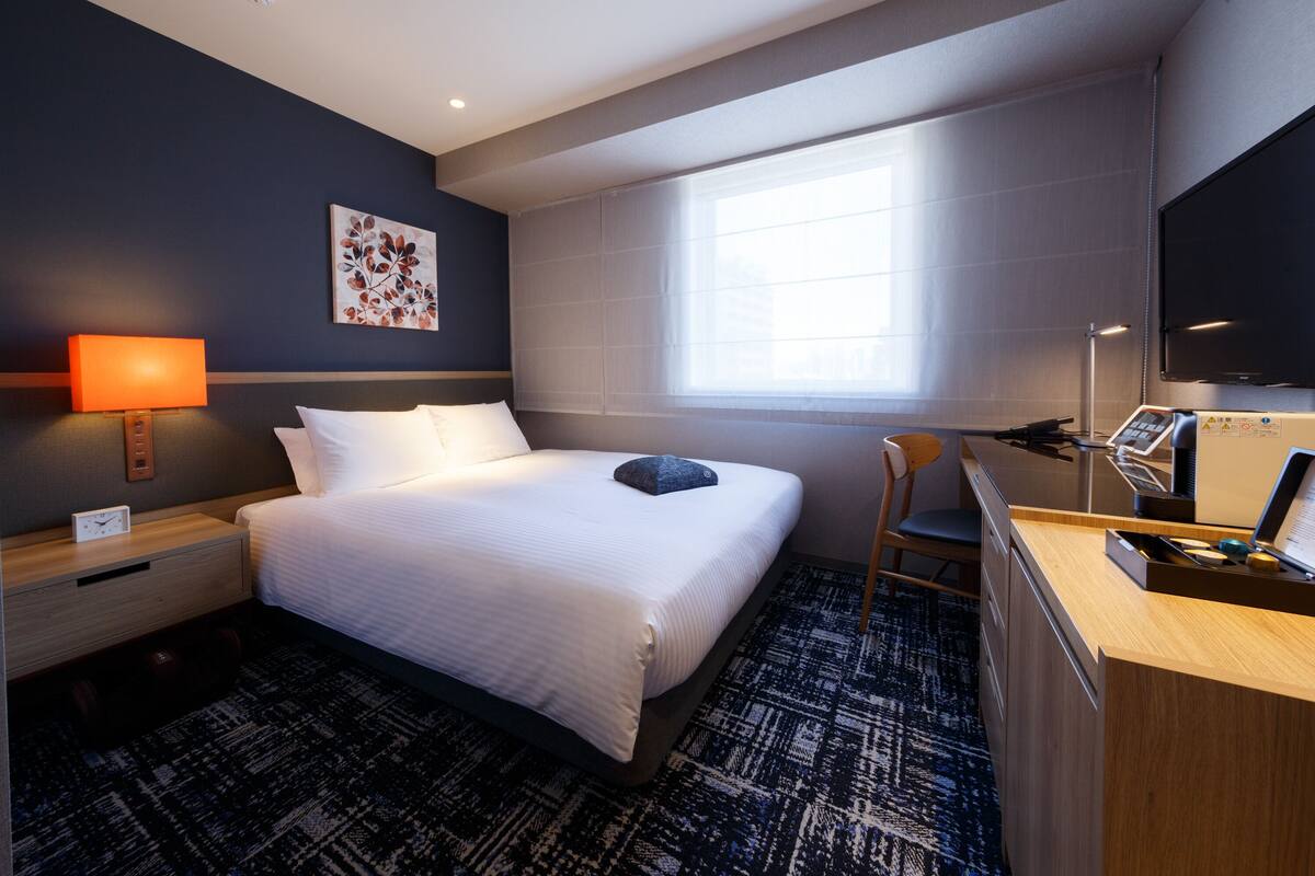 A hotel room with a bed, a desk, a chair, a TV, and a window.