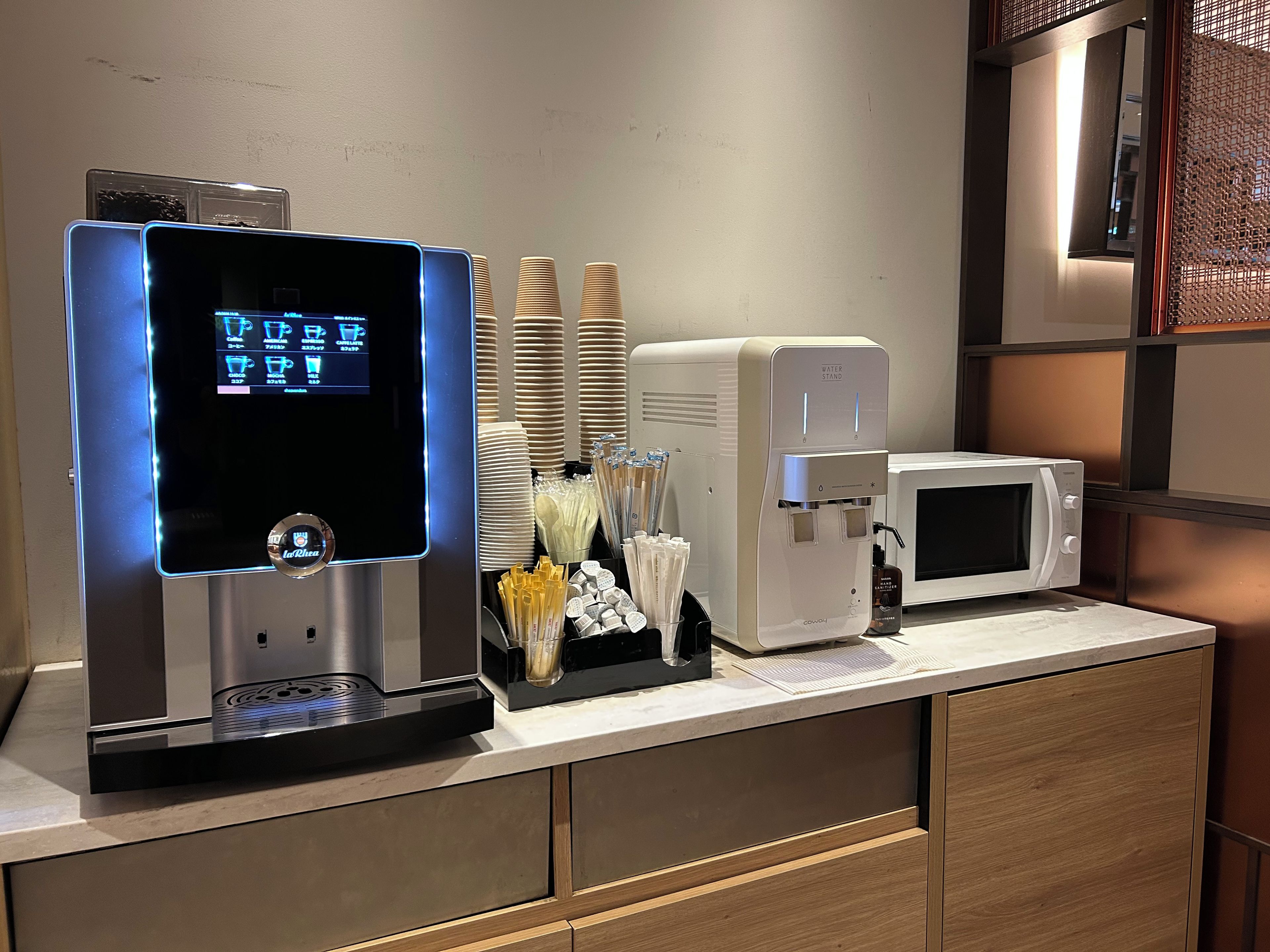 A self-service beverage station with a digital display, a water dispenser, and a microwave on a countertop.