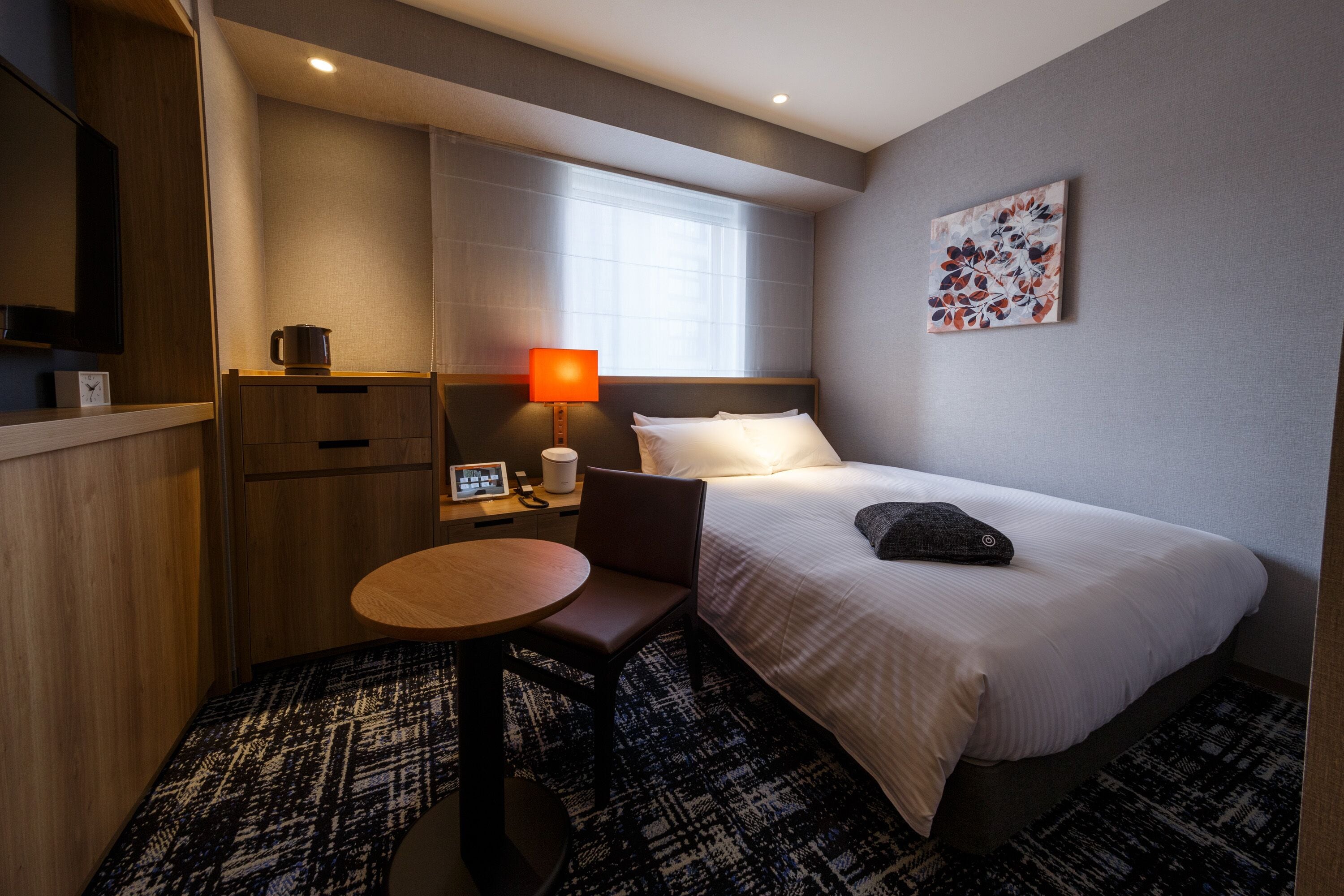 Une chambre d’hôtel avec un lit, un bureau et une chaise, une lampe, une horloge et un tableau au mur.
