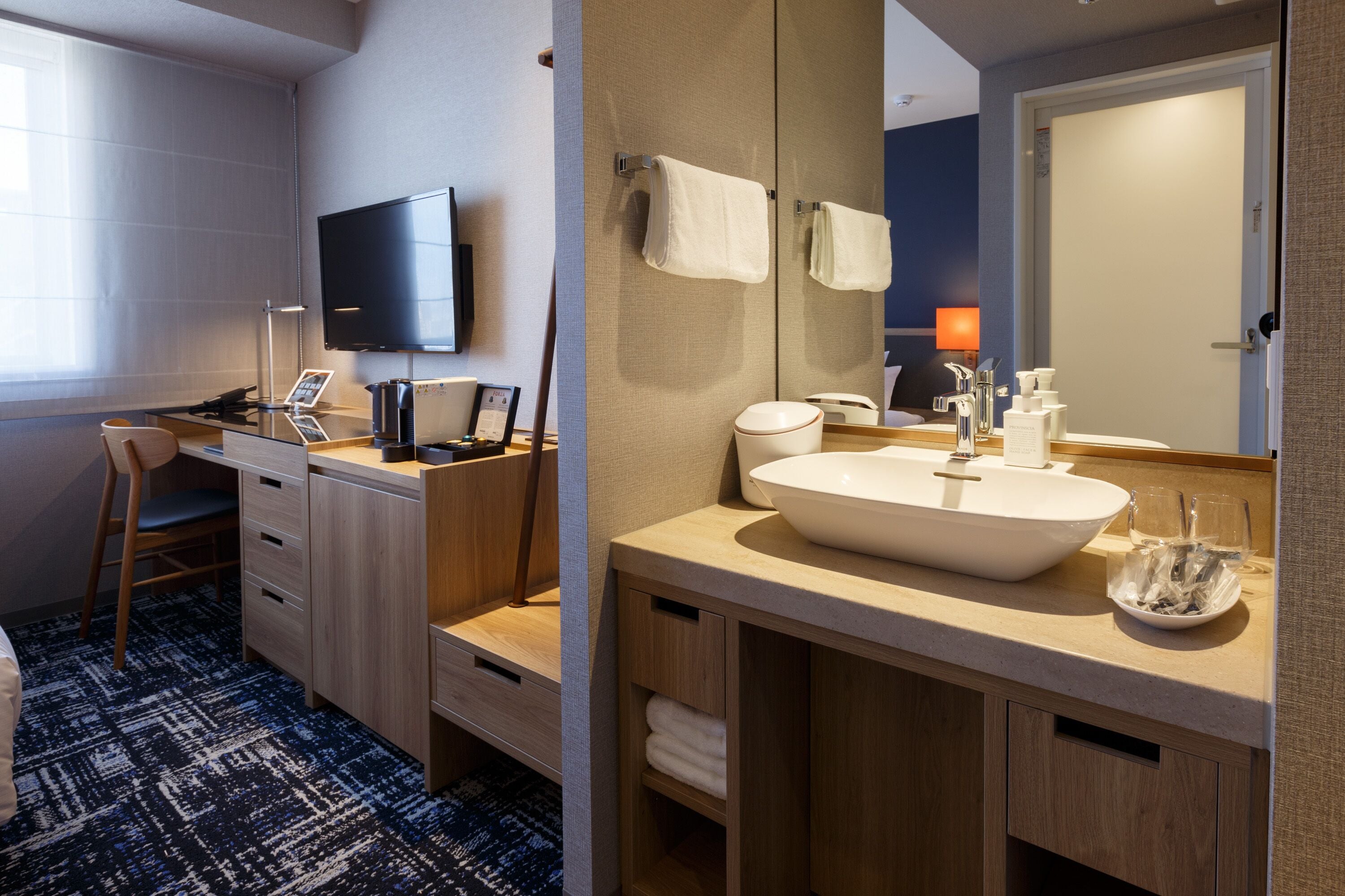 A modern hotel room with a large bed, a desk with a computer, a flat-screen TV, a large mirror, a sink, and a window with curtains.