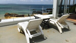 Un balcón con sillas de plástico blanco y una mesa, con vistas a un litoral rocoso y al mar.