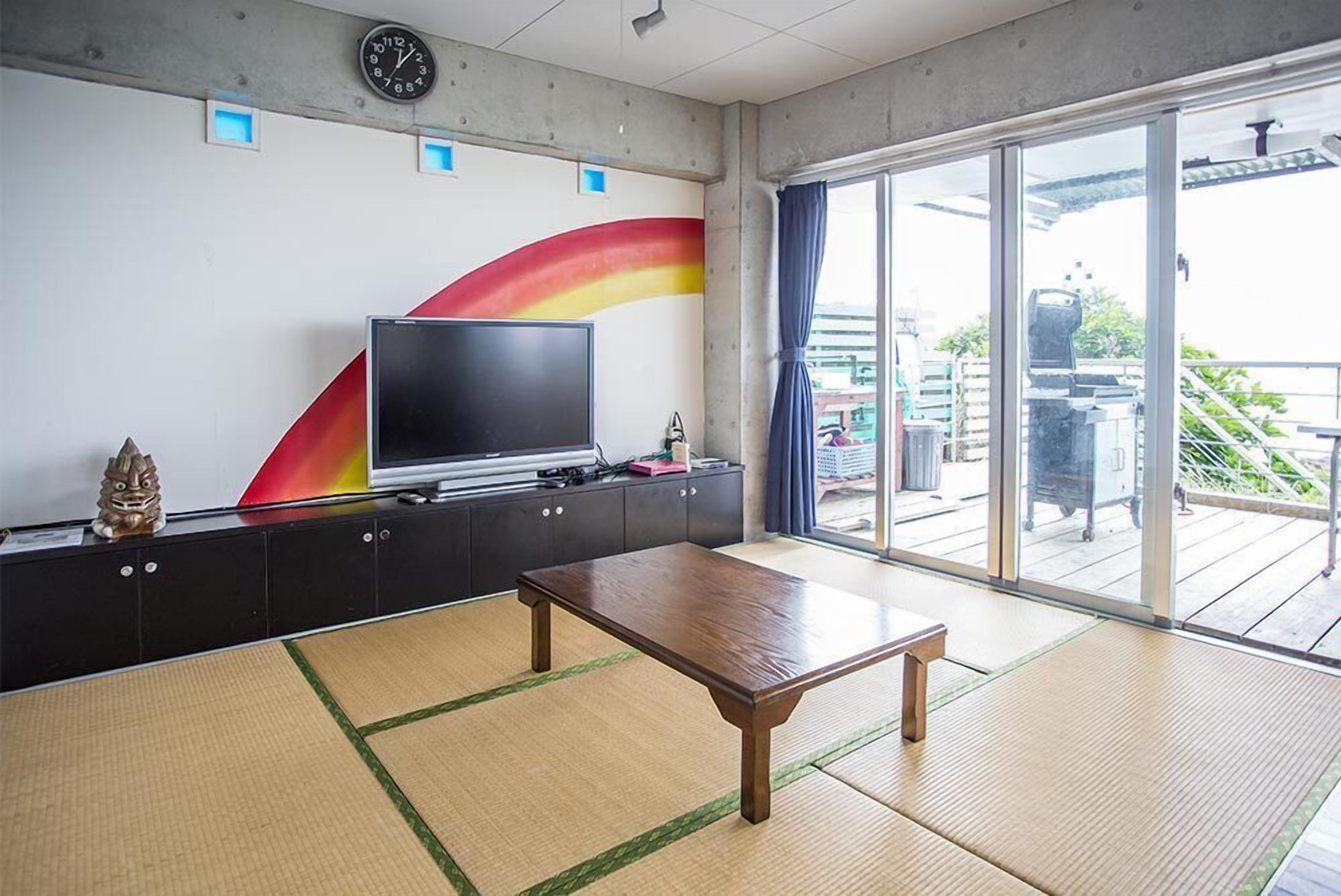 Une pièce avec des tatamis, un téléviseur posé sur un meuble, une horloge au mur et une vue sur le balcon extérieur.