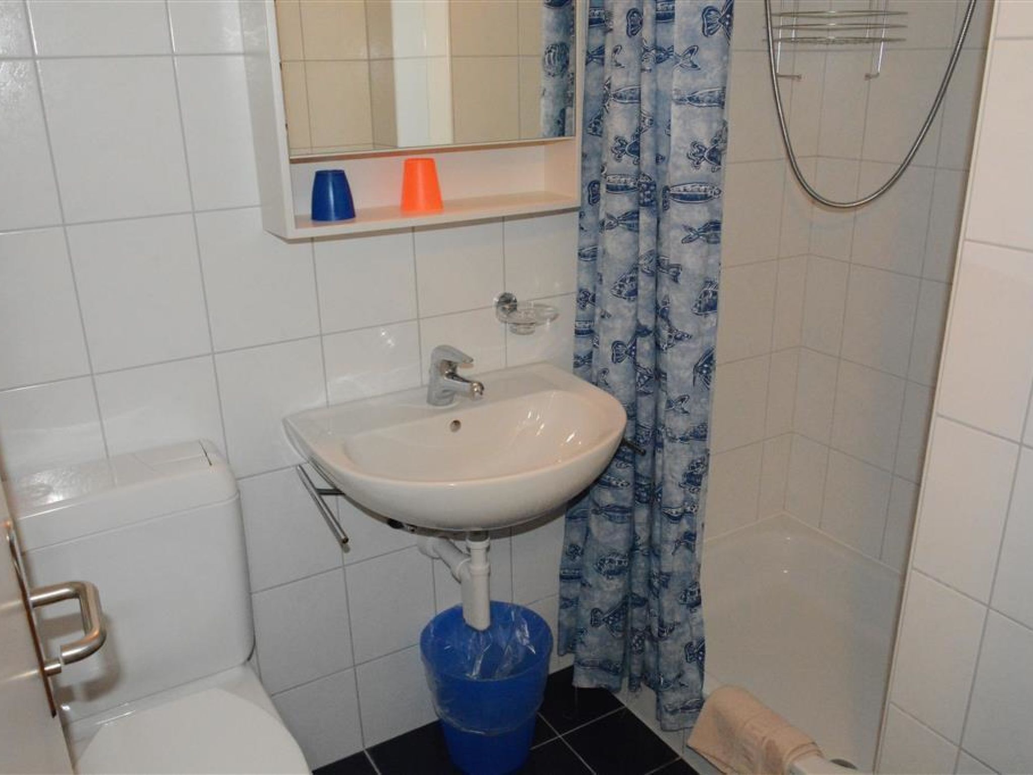 A bathroom with a white sink, a mirror above it, a blue bucket, and a shower curtain with a fish pattern.