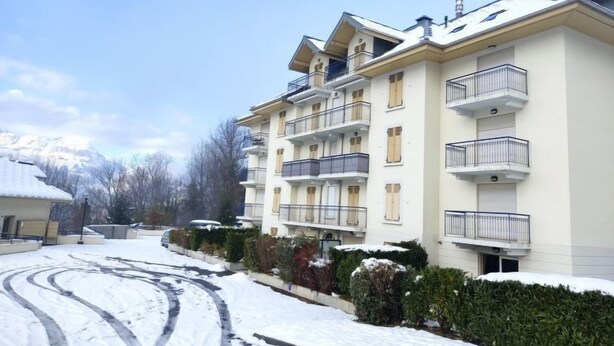 A multi-story residential building with balconies, a snow-covered driveway, and a mountainous backdrop.