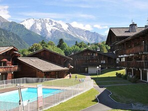 En træchalet med swimmingpool, omgivet af frodigt grønt og bjerge i baggrunden.