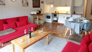 Un ambiente compatto con un divano rosso, un tavolino in legno e un'area pranzo con tavolo apparecchiato per quattro. In secondo piano, si intravede una cucina con pensili bianchi e un fornello.