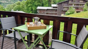 Un terrazzo in legno con un tavolo e due sedie, un vassoio con una caffettiera e tazze, e vista su una montagna e degli edifici sullo sfondo.