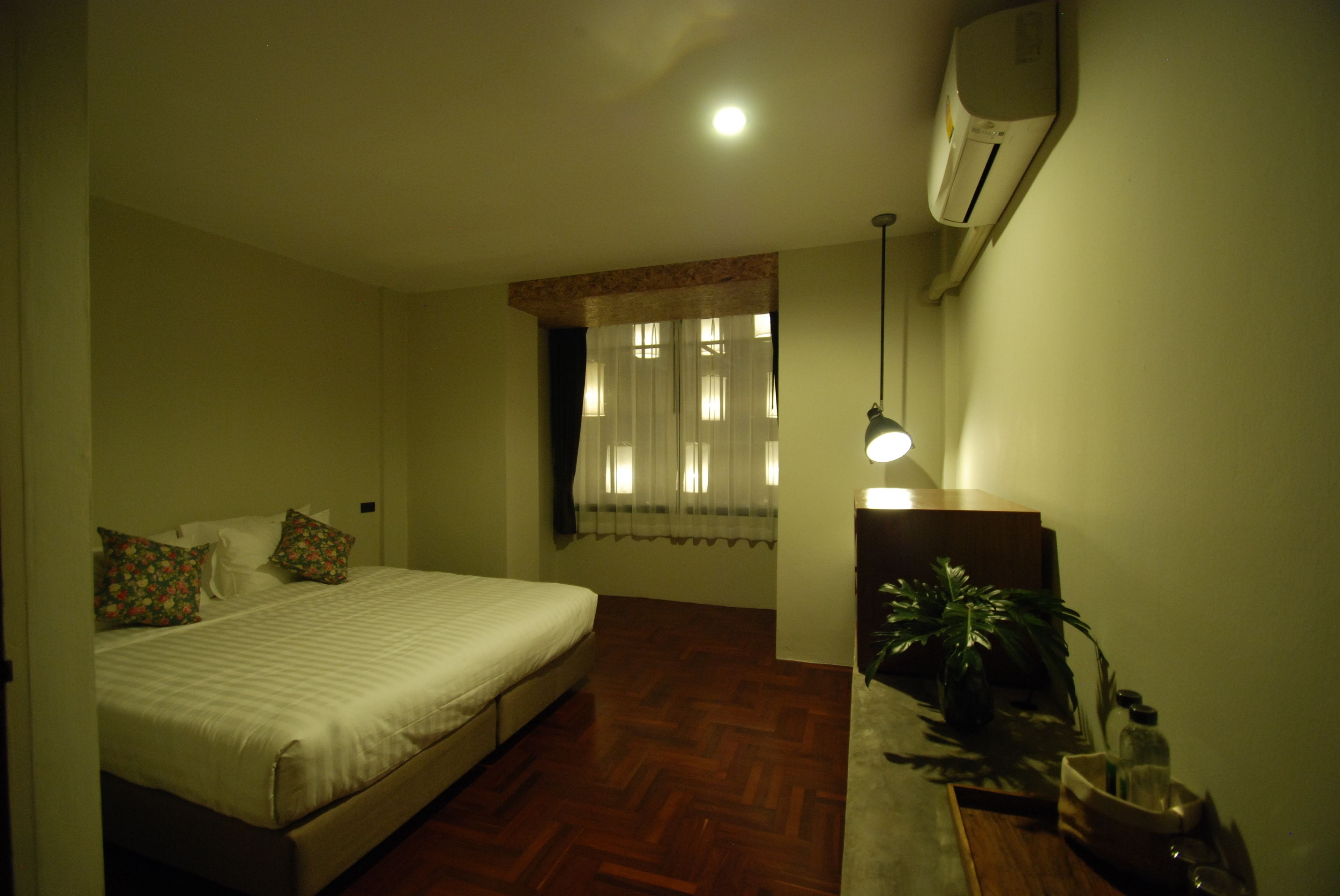 A bedroom with a bed, a desk with a lamp, a potted plant, and an air conditioning unit.