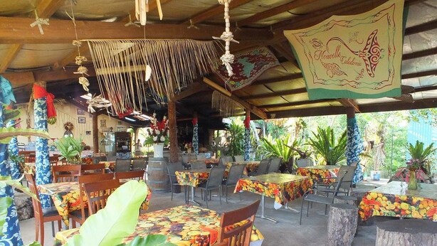 An outdoor restaurant with wooden tables and chairs, covered by a wooden roof, and decorated with hanging ornaments and a banner. - Fenepaza (Lifou)