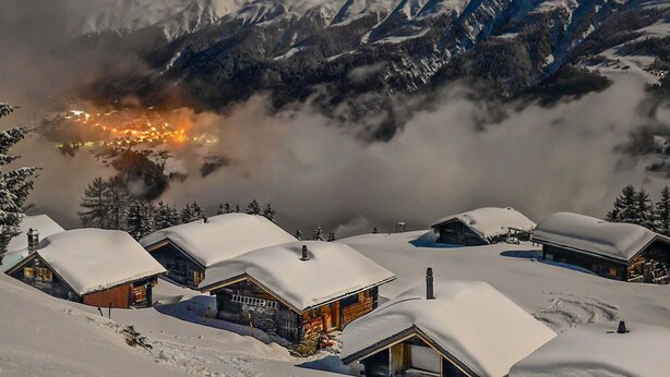 A snow-covered mountain village with wooden cabins, a visible fire, and a view of snow-capped peaks. - Alphütte Bielerhüs by Interhome (Fiesch)