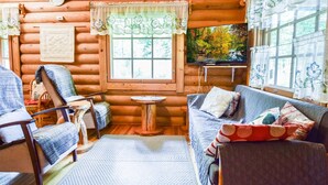 Interior acogedor de una cabaña de madera con suelo de madera, un sofá, un sillón, una mesita y una ventana con vistas a árboles.