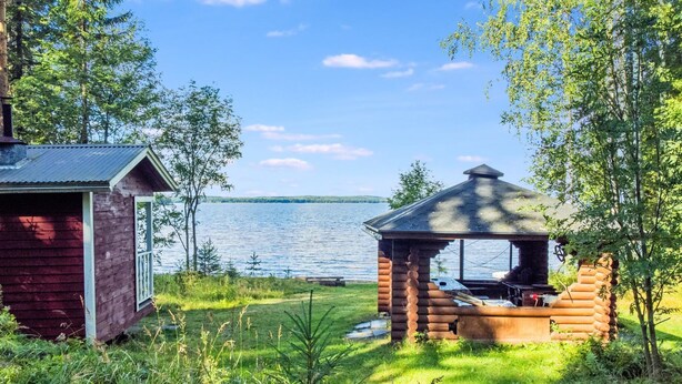 Una cabaña con un porche pequeño y una zona de parrilla, rodeada de árboles y un lago.