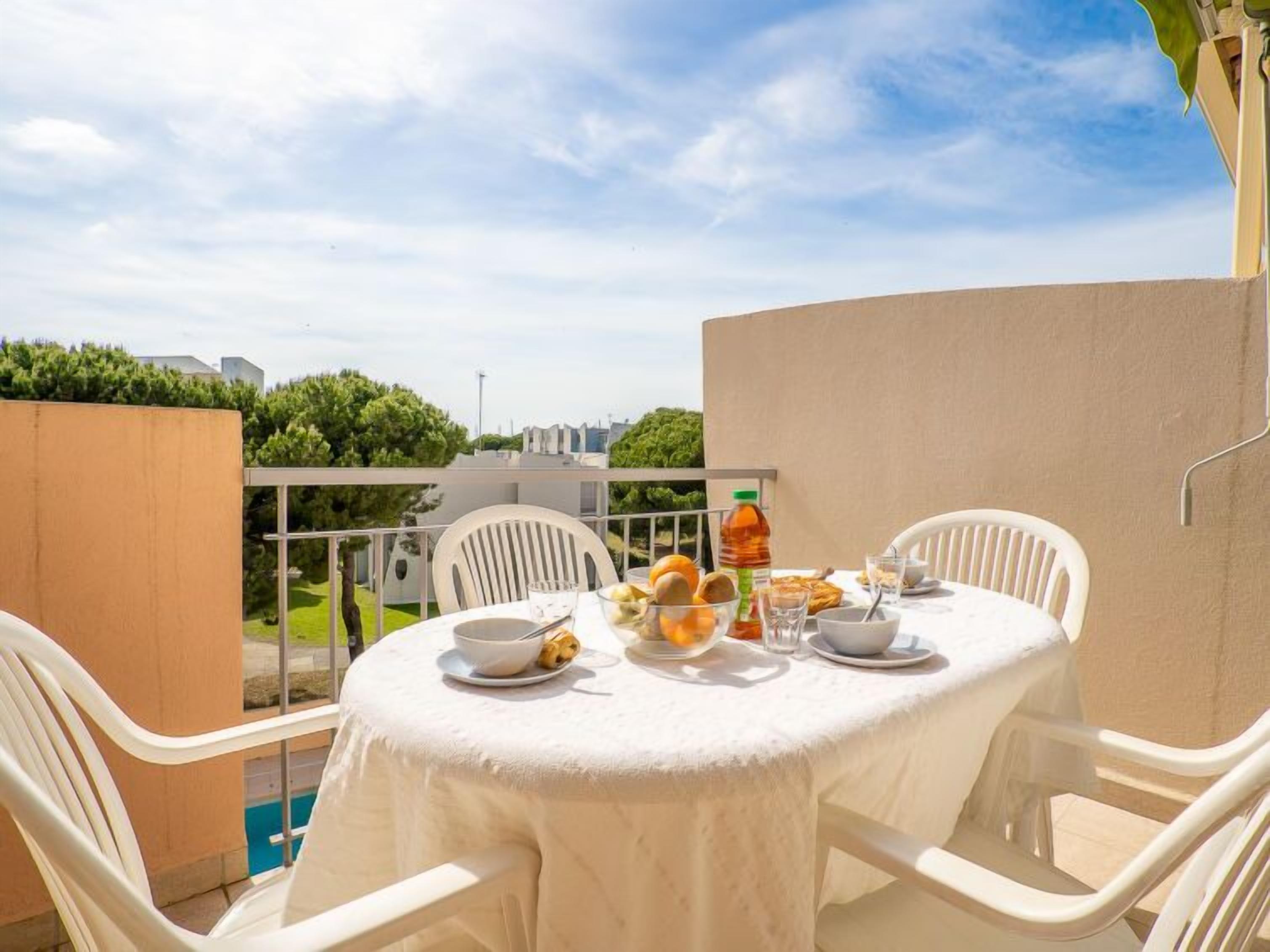 Een ronde tafel met een witte tafelkleed, omringd door witte stoelen, op een balkon met een helder uitzicht op de lucht en groen op de achtergrond.