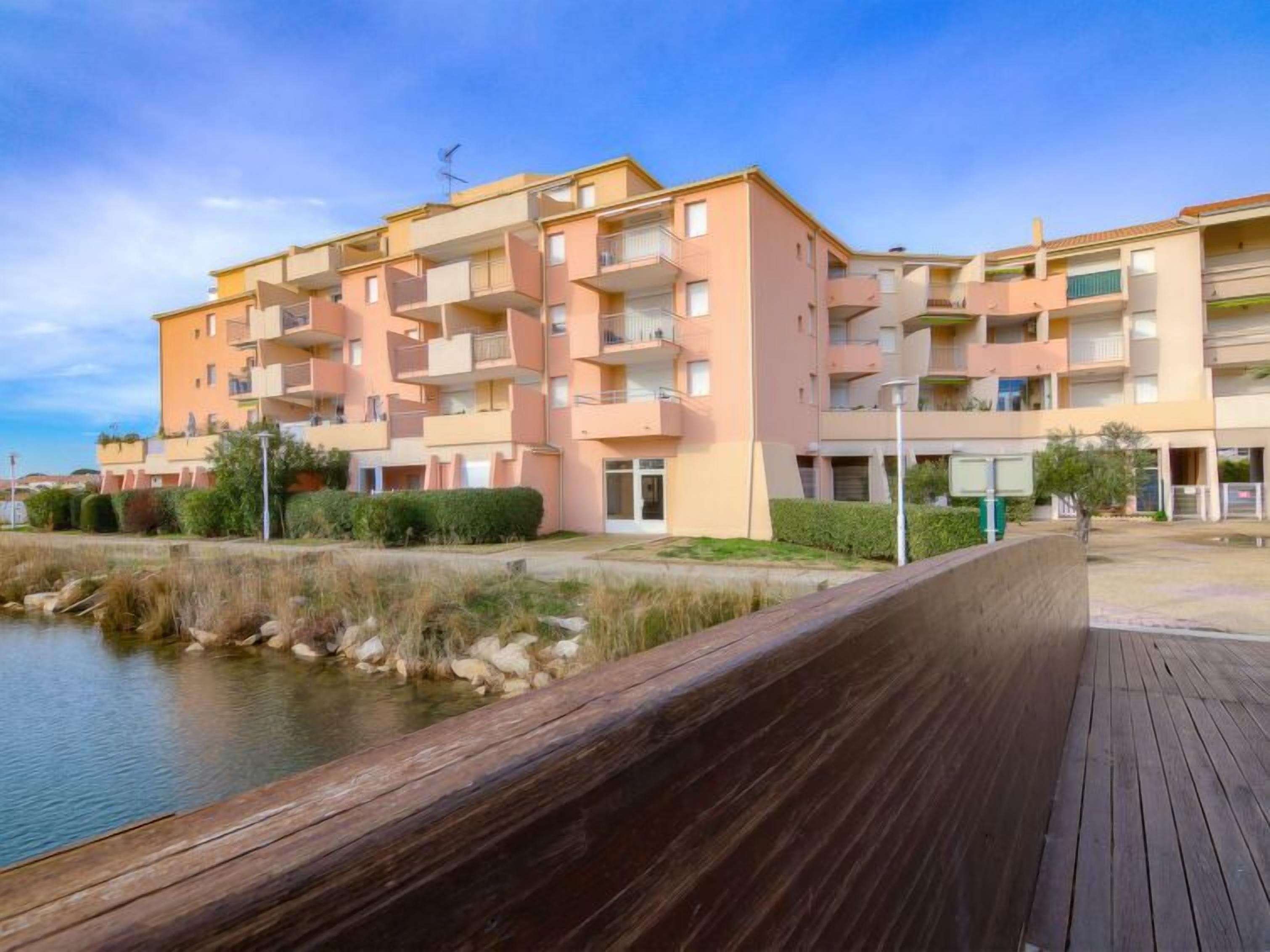 Un immeuble résidentiel à plusieurs étages, doté de balcons, d’une promenade en bois et d’une étendue d’eau au premier plan.