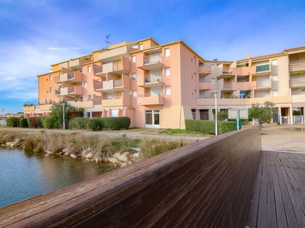 A multi-story residential building with balconies, a wooden boardwalk, and a body of water in the foreground.