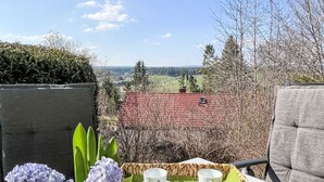 Una mesa con una bandeja de tazas blancas, platillos y una manzana verde, un florero de vidrio con flores moradas, y una vista de un edificio de tejados rojos y árboles.