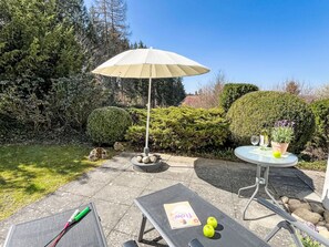 A patio with a table, a bench, and an umbrella. There are trees and bushes in the background.