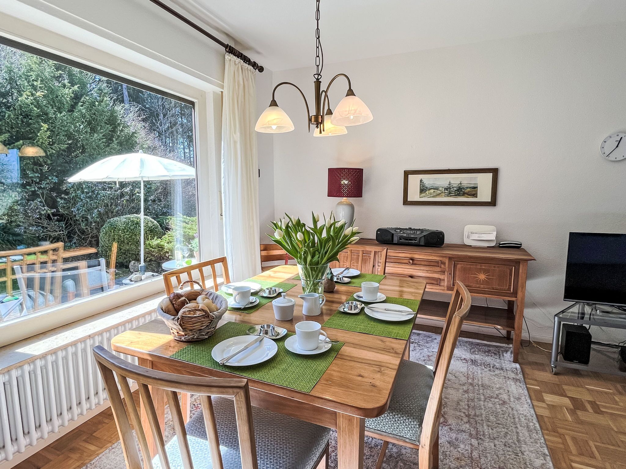 A dining area with a table set for four, a radio, a clock, and a window with a view of greenery.