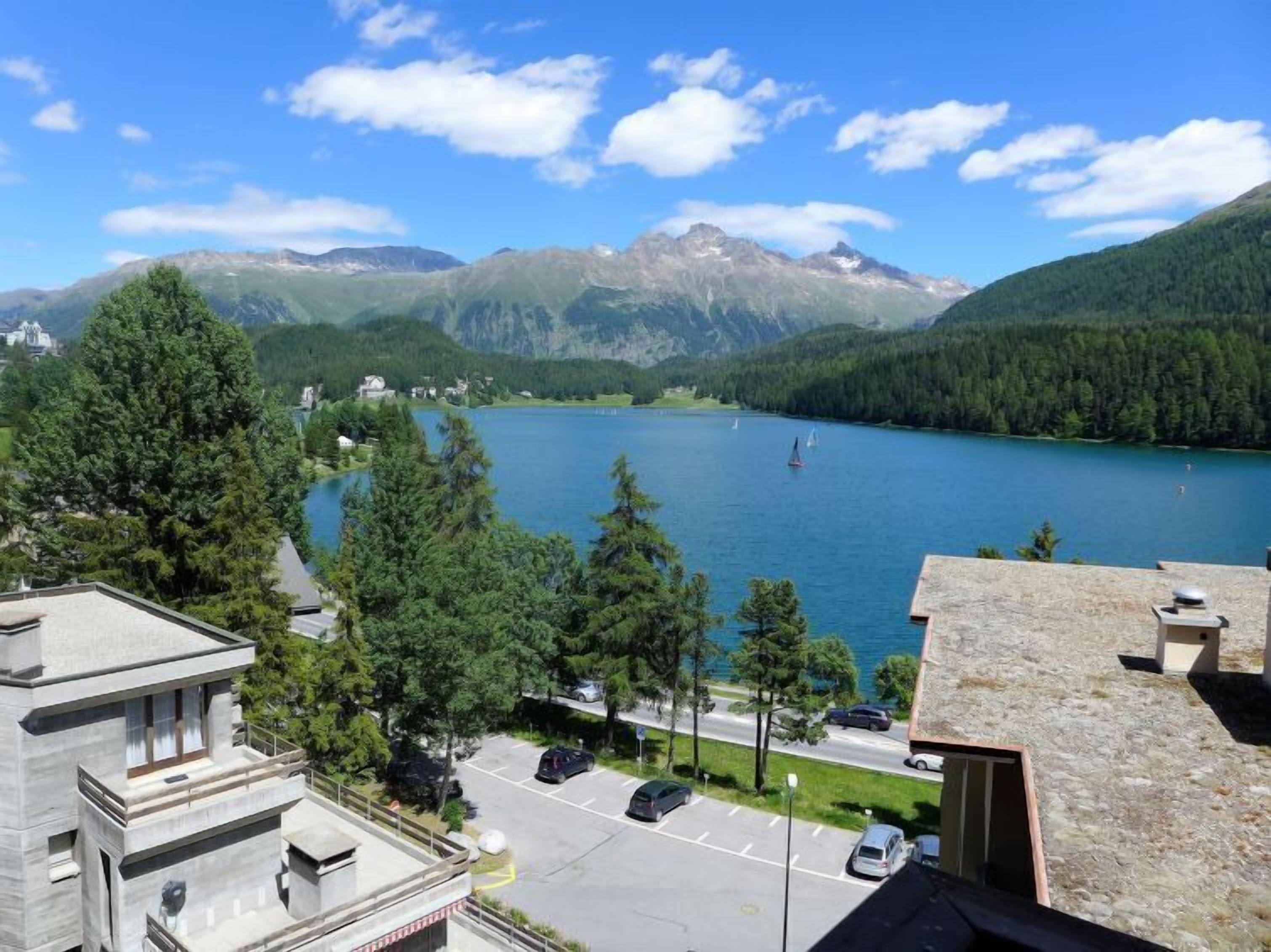 A view of a lake surrounded by mountains, with a parking area and buildings in the foreground.