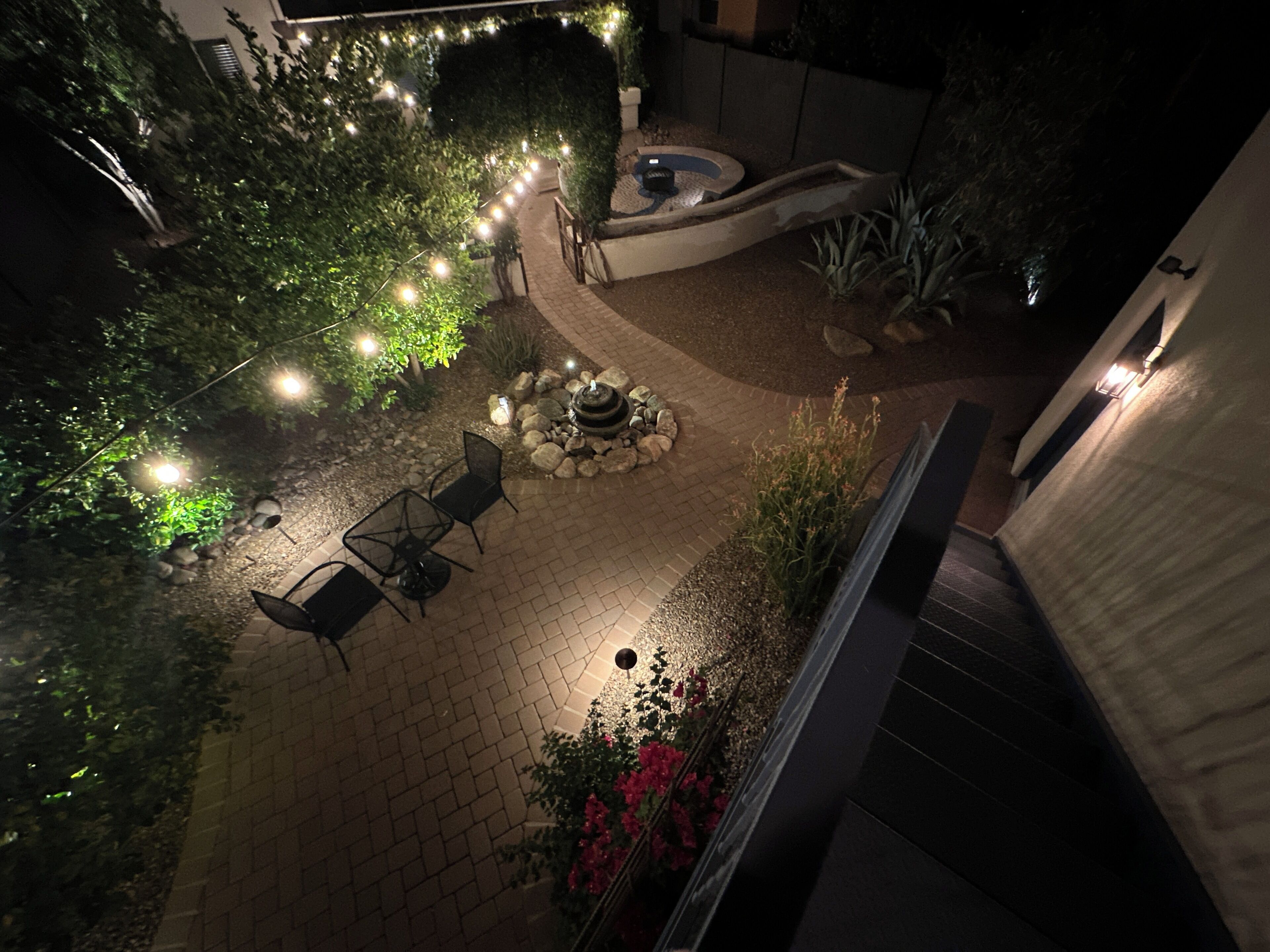 An outdoor patio area with a brick floor, a seating arrangement with chairs and a table, a small water feature, and string lights.