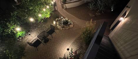 An outdoor patio area with a brick floor, a seating arrangement with chairs and a table, a small water feature, and string lights.