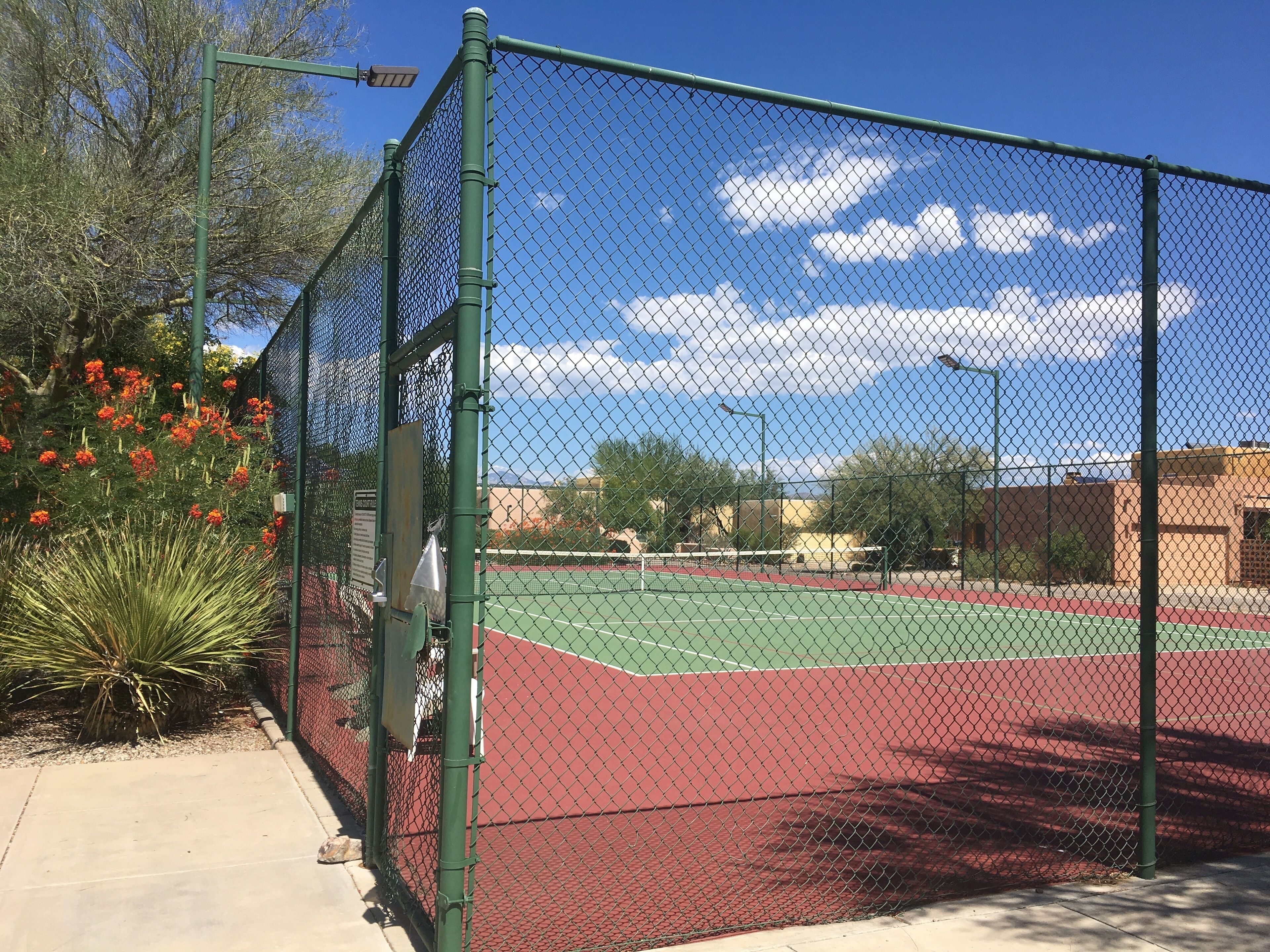 A tennis court enclosed by a chain-link fence with a red surface, surrounded by trees and a clear sky.