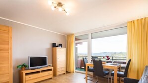 Ein modernes Wohnzimmer mit Flachbildfernseher, Holzmöbeln, einer Essecke mit Tisch und Stühlen sowie einem Balkon mit gelben Vorhängen.