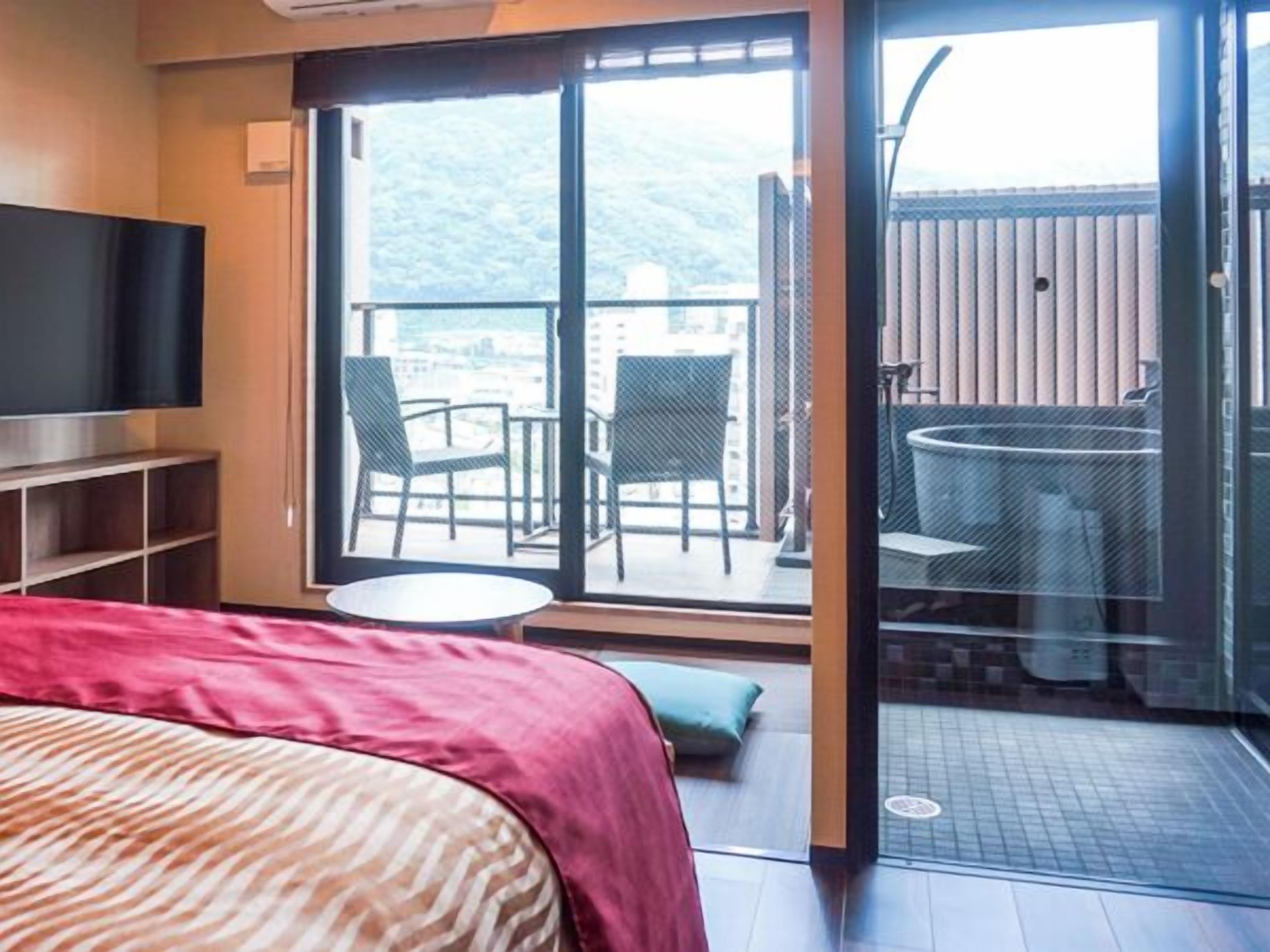 Et hotelværelse med seng, fjernsyn, balkon med stole og bord samt udsigt til det fri.