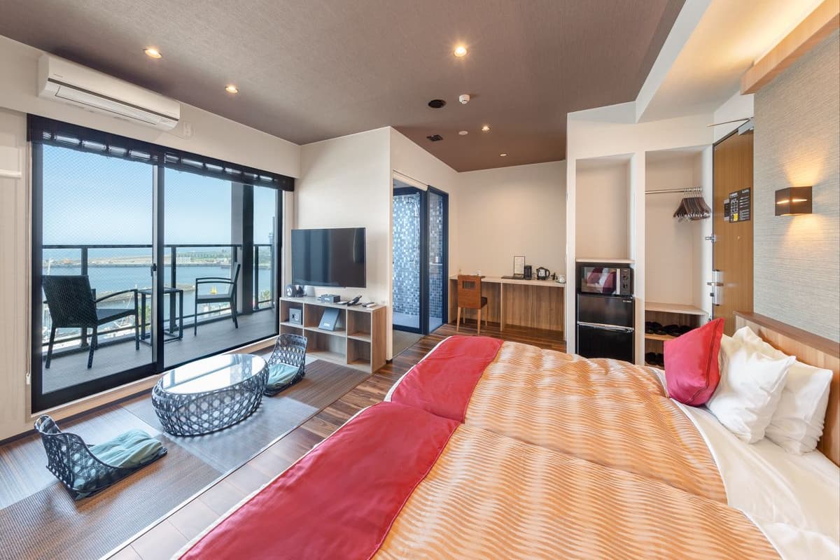 A modern hotel room with a large bed, a TV, a desk, and a balcony with a view of the sea.