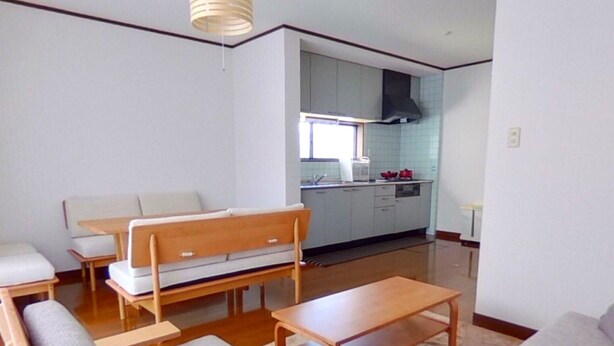 A compact living space with a sofa, a wooden bench, and a dining table. A kitchen area is visible in the background with cabinets and a stove. - Wakayama House No.7 (Wakayama)