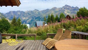 Um deck de madeira com cadeiras de vime e uma mesa, com vista para uma paisagem montanhosa com uma cabana.