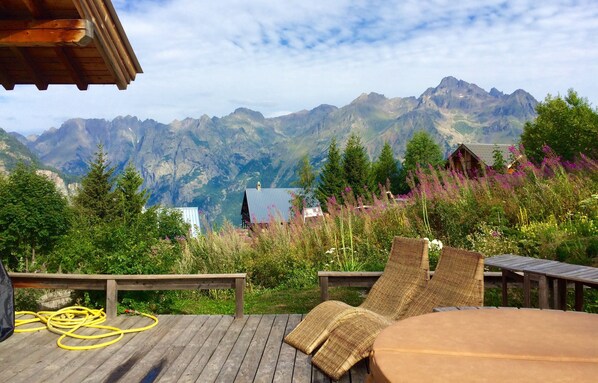 A wooden deck with wicker chairs and a table, overlooking a mountainous landscape with a cabin.