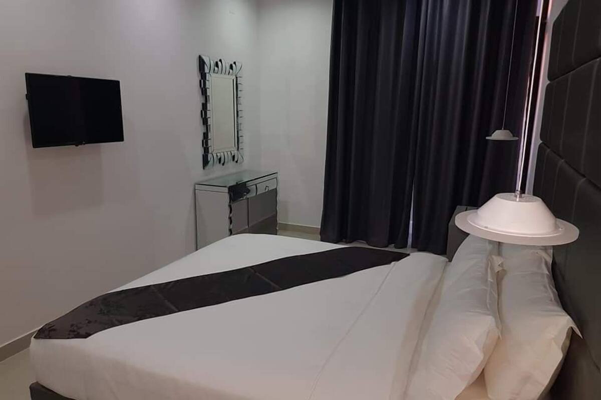 A neatly made bed with white and black bedding, a bedside table, a mirror, and a TV mounted on the wall.