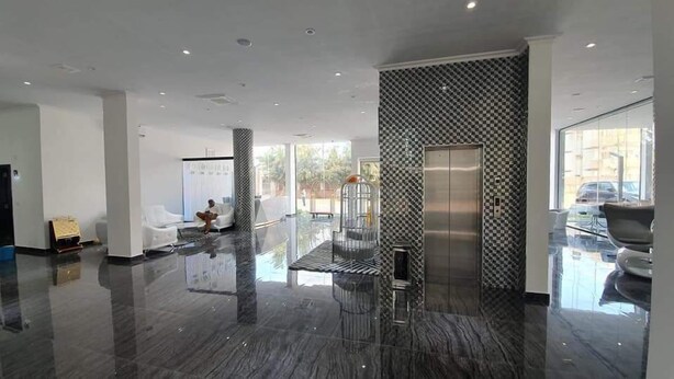 A spacious lobby with a modern design, featuring a glass elevator, a seating area, and a large window offering a view of the outdoors.