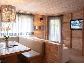 A modern living room with a wooden ceiling, a glass-top dining table with a bench, a sofa, and a TV mounted on the wall.