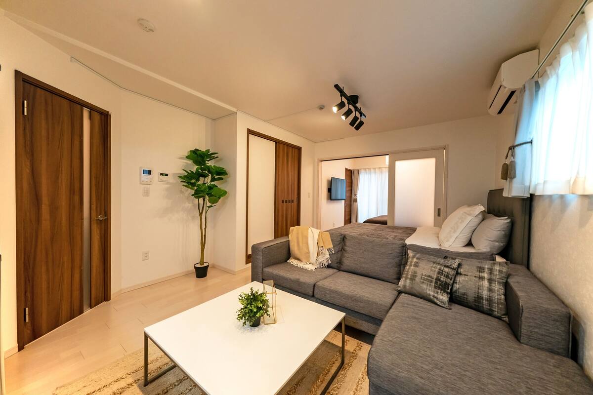 A modern living room with a grey sofa, a white coffee table, and a potted plant. There is a wooden door and a wall-mounted air conditioner.