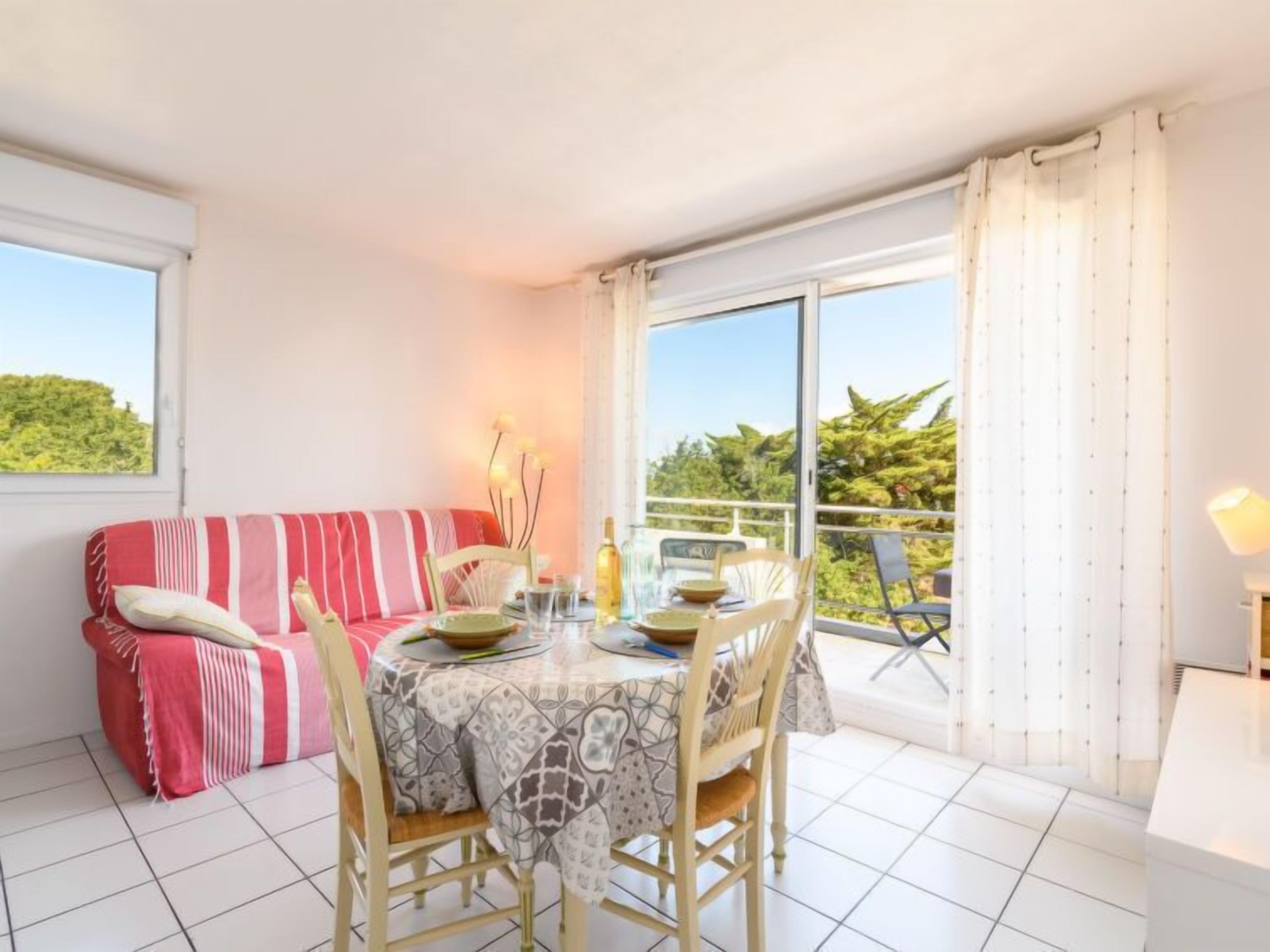Un coin repas avec une table dressée pour deux personnes, un canapé à rayures rouges et un balcon offrant une vue sur la verdure.