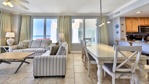 Un vaste espace de vie comprenant un canapé, une table à manger et des chaises, de grandes fenêtres avec des rideaux, un ventilateur de plafond et un coin cuisine avec des meubles en bois et des appareils électroménagers.