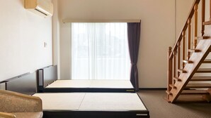 Een kamer met een futonbed, een houten stoel, een klein tafeltje, een houten trap en een groot raam met gordijnen.