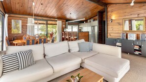 A modern cabin interior with a wooden ceiling, a sectional sofa, a dining area, and a kitchen. - Sensational Sun Trap on Estuary Frontage - Pauanui (Pauanui)