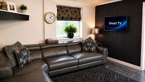 Una sala moderna con sofá de cuero, cojines decorativos, un reloj de pared, una planta en maceta, un televisor inteligente y una ventana con persianas.
