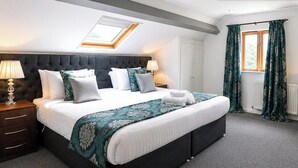Quarto de hotel com cama, escrivaninha, cadeira, abajur, janela com cortinas e claraboia.