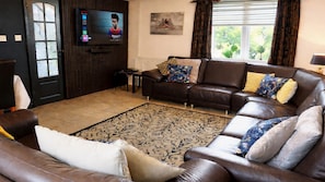 Ein Wohnzimmer mit braunem Ledersofa, einem gemusterten Teppich, einem an der Wand befestigten Fernseher und einem Fenster mit Vorhängen.