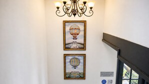 Um corredor com um candelabro, duas obras de arte emolduradas retratando balões de ar quente, e uma porta com um aviso sobre carregamento de veículos elétricos.