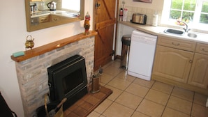 A kitchen with a stone fireplace, wooden cabinets, a dishwasher, and a window with a view of greenery. - Macbeth Cottage (Stratford-upon-Avon)