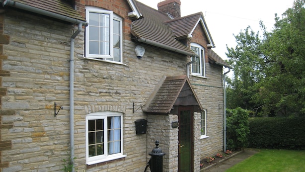 A stone house with a gabled roof, a front door, and a small garden with flowers. - Macbeth Cottage (Stratford-upon-Avon)