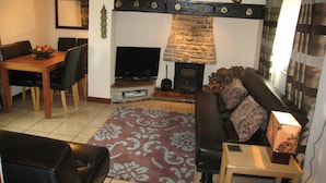 A cozy living room with a stone fireplace, a television, a dining table with chairs, and a patterned rug. - Macbeth Cottage (Stratford-upon-Avon)