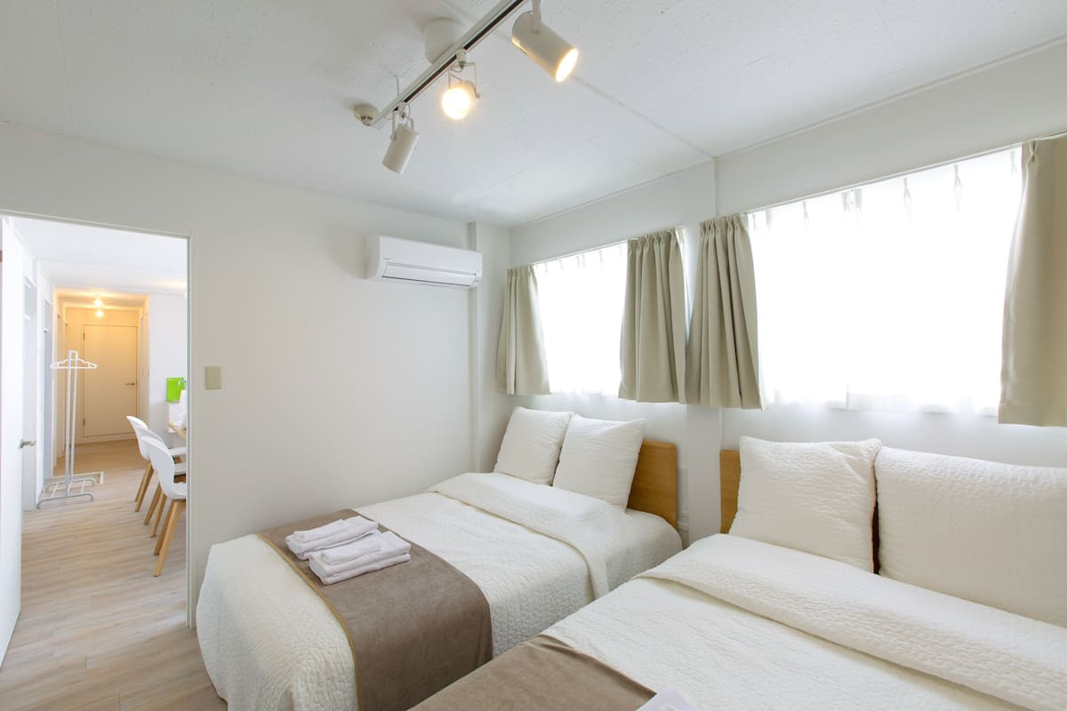 A hotel room with two beds, a desk, and a chair. There is an air conditioning unit on the wall and curtains on the windows.