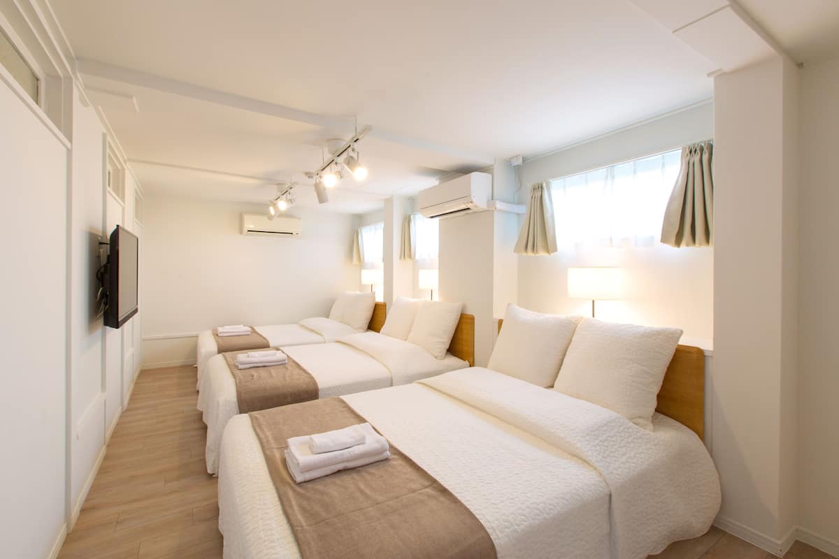 A hotel room with three beds, each with a brown blanket and white pillows. There are ceiling lights, an air conditioning unit, and a TV mounted on the wall.